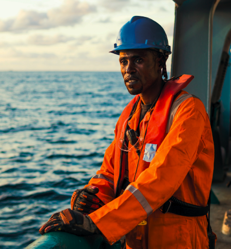Man with a worried expression, wearing a blue hard hat and orange overalls, on deck looking out to sea.